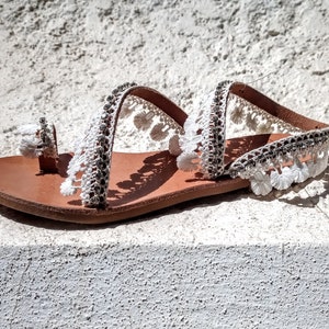 White leather sandals with white pom-pom trim and a brown leather sole.