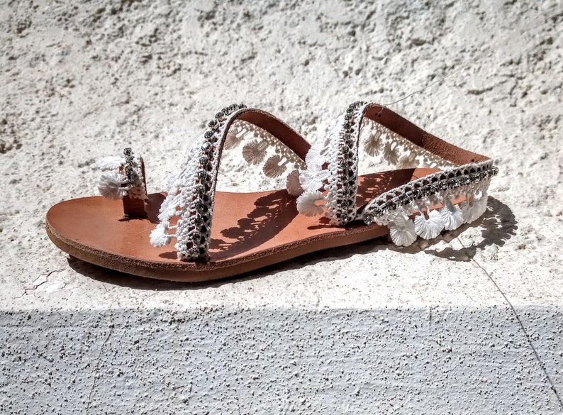White leather sandals with white pom-pom trim and a brown leather sole.