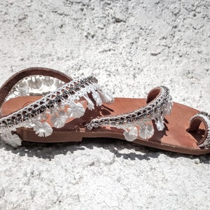 White leather sandals with white lace trim and rhinestones. The sandals have a toe loop and an ankle strap.