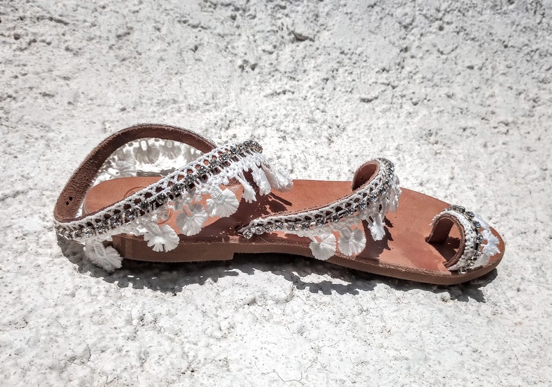 White leather sandals with white lace trim and rhinestones. The sandals have a toe loop and an ankle strap.