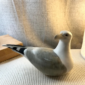 May include: A ceramic figurine of a seagull with a white head and neck, featuring soft grey shading on the wings and body. The bird has a light-colored beak, a dark eye, and black markings on the tips of its tail feathers, captured in a resting pose.