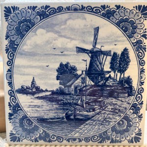May include: A square ceramic tile featuring a circular blue and white illustration of a Dutch landscape. The scene depicts a windmill, a small boat docked at a wooden pier, and a distant church tower, all framed by a decorative floral border.