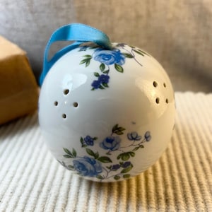 May include: A spherical white ceramic pomander featuring a blue floral pattern with green leaves and a light blue ribbon loop at the top. The surface includes small perforated holes arranged in clusters for scent diffusion.