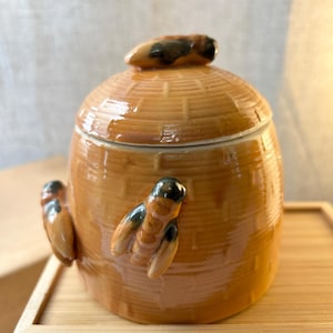 May include: A glossy, honey-colored ceramic jar shaped like a traditional beehive, featuring a textured, woven-look surface. The jar includes a matching lid with a sculpted bee on top and two additional sculpted bees attached to the sides.