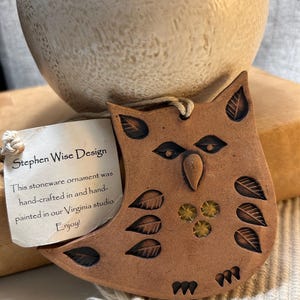 May include: A brown stoneware owl ornament with leaf-shaped indentations and three yellow glass accents. A white tag attached with string reads: Stephen Wise Design. This stoneware ornament was hand-crafted in and hand-painted in our Virginia studio. Enjoy!