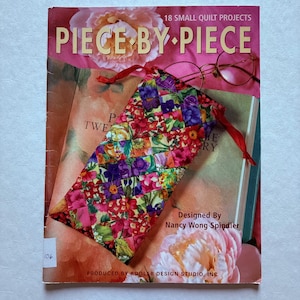 May include: A book titled "Piece by Piece" with a floral design, featuring a small, colorful quilted bag with a red drawstring. The book cover displays text and a peony illustration. A pair of glasses rests on the book.