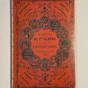 May include: A vintage red booklet with black ornate designs and text. The cover features the words "PATTERN'S FOR WORKS" and "MEDAILLES DE 1ERE CLASSE". Circular illustrations surround the central text. The booklet's theme is related to exhibitions.