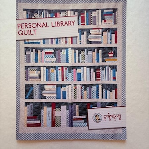 May include: A quilted wall hanging depicting a bookshelf filled with colorful books. The words "PERSONAL LIBRARY QUILT" are displayed across the top. The quilt has a gray border with a blue and white pattern and a signature that says "crimson tate".