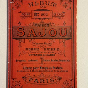 May include: A vintage red and black album cover with the text "ALBUM DE POINT R: 902 DE CROIX MAISON SAJOU". The cover also includes text for "BRODERIES TAPISSERIES" and "PARIS". The design features ornate floral and geometric patterns.
