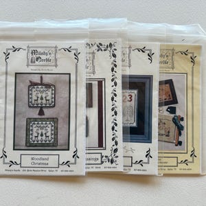 May include: Four cross-stitch pattern packets in clear plastic sleeves. The packets feature various designs, including a Christmas theme with deer and the text "Woodland Christmas." Other packets show framed images and the text "Blessings."