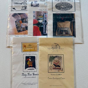 May include: A collection of primitive needlepunch patterns from Notforgotten Farm, featuring various designs. The patterns include images of a turkey, tulips, a meadow scene, a Santa Claus, and a crow. Each pattern is packaged in a clear plastic sleeve.