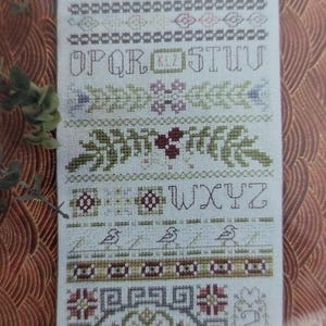 May include: A cross-stitch sampler with embroidered letters, floral patterns, and bird motifs. The design includes the alphabet, decorative borders, and a central circular pattern. The color palette features shades of green, pink, and blue on a white background.