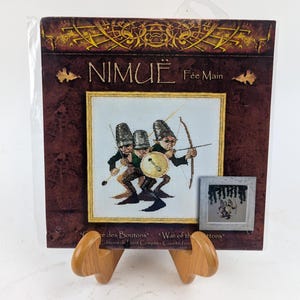May include: A cross-stitch pattern featuring three whimsical fairies with pointed hats, holding a shield and bow and arrow. The pattern is titled "Nimue Fée Main" and is part of the "War of the Buttons" collection.