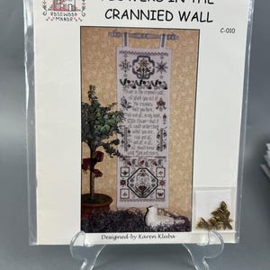 May include: A white cross-stitch pattern with a floral design and the text "Flowers in the Crannied Wall" and "Tourer in the crammed wall el pluck you out of The cranes fold you fiere, Troof and all, in my fand, Flower-but if e could understood what you are, Prod and di should koow wid "Dod and man o". The pattern is framed and displayed on a stand.