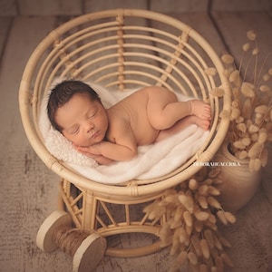 Op de afbeelding: Een pasgeboren baby slaapt rustig in een rieten stoel met een wit deken. De stoel staat op een houten vloer met gedroogde bloemen in een vaas ernaast.