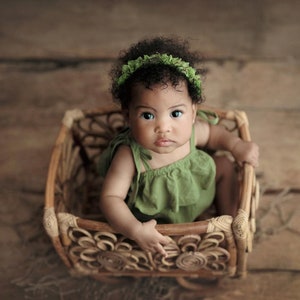 Op de afbeelding: Een babymeisje in een groene jurk en een groene bloemenkrans zit in een geweven rieten mand. De mand heeft een decoratief bloemenpatroon.