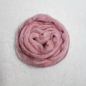May include: A round bundle of pink fiber with shimmering threads. The fiber is tightly wound, creating a textured surface. The color is a blend of light and dark pink hues, with the shimmering threads adding a touch of sparkle. Ideal for crafting.