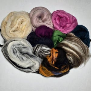 May include: A collection of colorful wool roving in a black handled tray. The wool is in various shades of cream, pink, purple, green, and brown. The tray is oval-shaped and has two black handles.