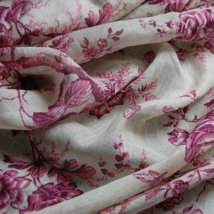 May include: A close-up of a white linen fabric with a pink floral pattern. The fabric is folded and draped, showing the intricate details of the design.