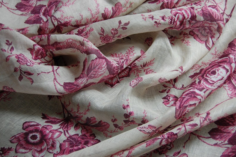 May include: A close-up of a white linen fabric with a pink floral pattern. The fabric is folded and draped, showing the intricate details of the design.