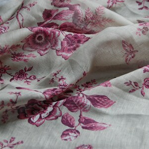 May include: A close-up of a sheer fabric with a floral pattern. The fabric is a light beige color with pink flowers and leaves. The fabric is draped and has a soft, flowing texture.