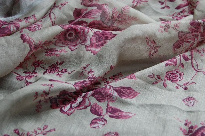 May include: A close-up of a sheer fabric with a floral pattern. The fabric is a light beige color with pink flowers and leaves. The fabric is draped and has a soft, flowing texture.
