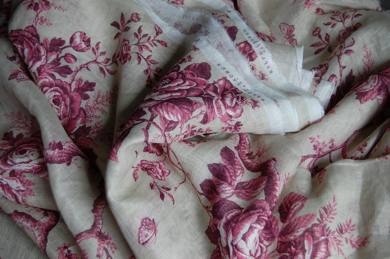 May include: A close-up of a beige linen fabric with a pink floral pattern. The fabric is folded and has a white label with the text "RALPH LAUREN" on it.
