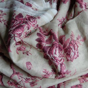 May include: A close-up of a beige linen fabric with a repeating pattern of pink roses and butterflies. The fabric is folded and draped, showing the texture and detail of the design.
