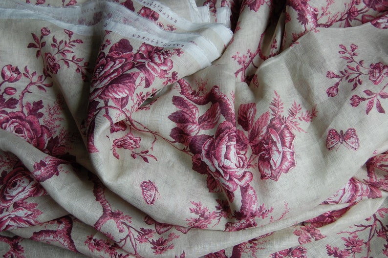 May include: A close-up of a beige linen fabric with a repeating pattern of pink roses and butterflies. The fabric is folded and draped, showing the texture and detail of the design.