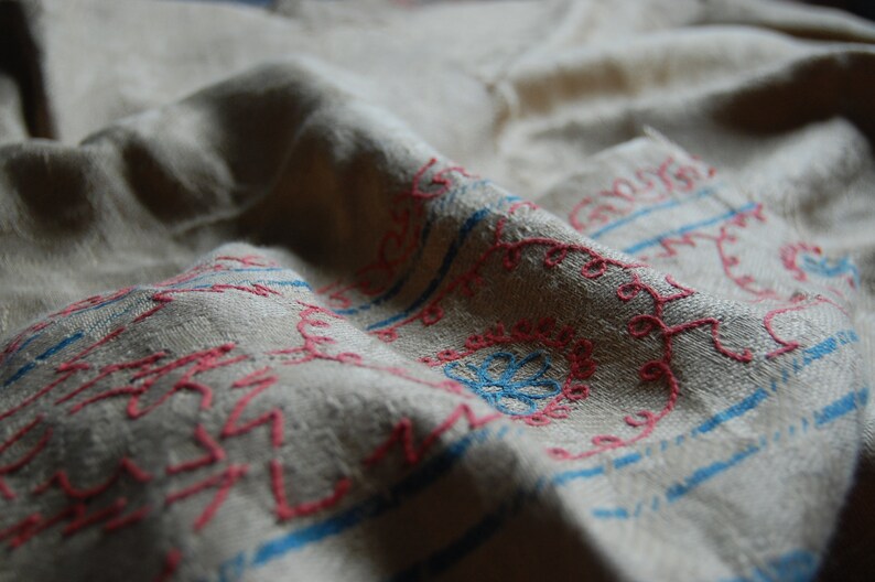 Puede incluir: Primer plano de una tela blanca con bordados rosas y azules. La tela est&aacute; doblada y tiene una superficie texturizada. El bordado es de patr&oacute;n floral.