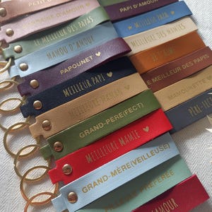 May include: A collection of rectangular faux leather keychains in various colours including red, blue, green, tan, and purple. Each keychain features a gold-toned metal ring, a gold rivet, and gold embossed French text such as Maman Je T'aime.