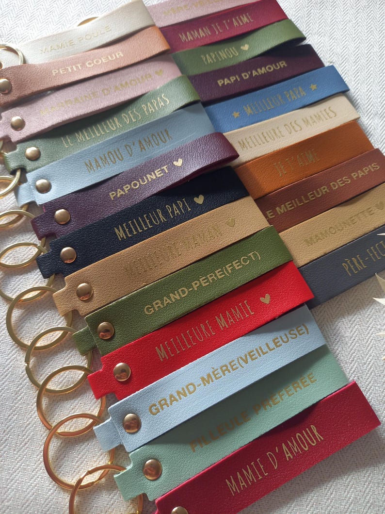 May include: A collection of rectangular faux leather keychains in various colours including red, blue, green, tan, and purple. Each keychain features a gold-toned metal ring, a gold rivet, and gold embossed French text such as Maman Je T'aime.