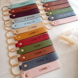 May include: A collection of rectangular faux leather keychains in various colours including light blue, brown, burgundy, grey, yellow, red, olive green, and pink. Each keychain features a gold-tone metal ring, a gold rivet, and custom text.