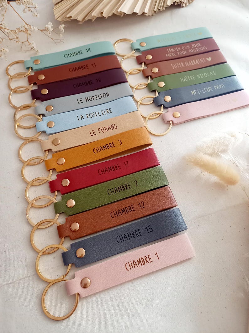 May include: A collection of rectangular faux leather keychains in various colours including light blue, brown, burgundy, grey, yellow, red, olive green, and pink. Each keychain features a gold-tone metal ring, a gold rivet, and custom text.