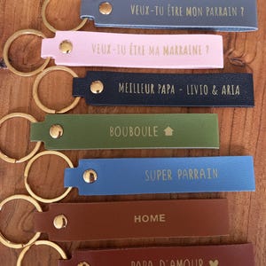 May include: Seven rectangular keychains in various colours including blue, pink, navy, green, light blue, and brown. Each features a gold metal ring, a gold rivet, and gold text including phrases like Veux-tu &ecirc;tre mon parrain, Meilleur Papa, and Home.