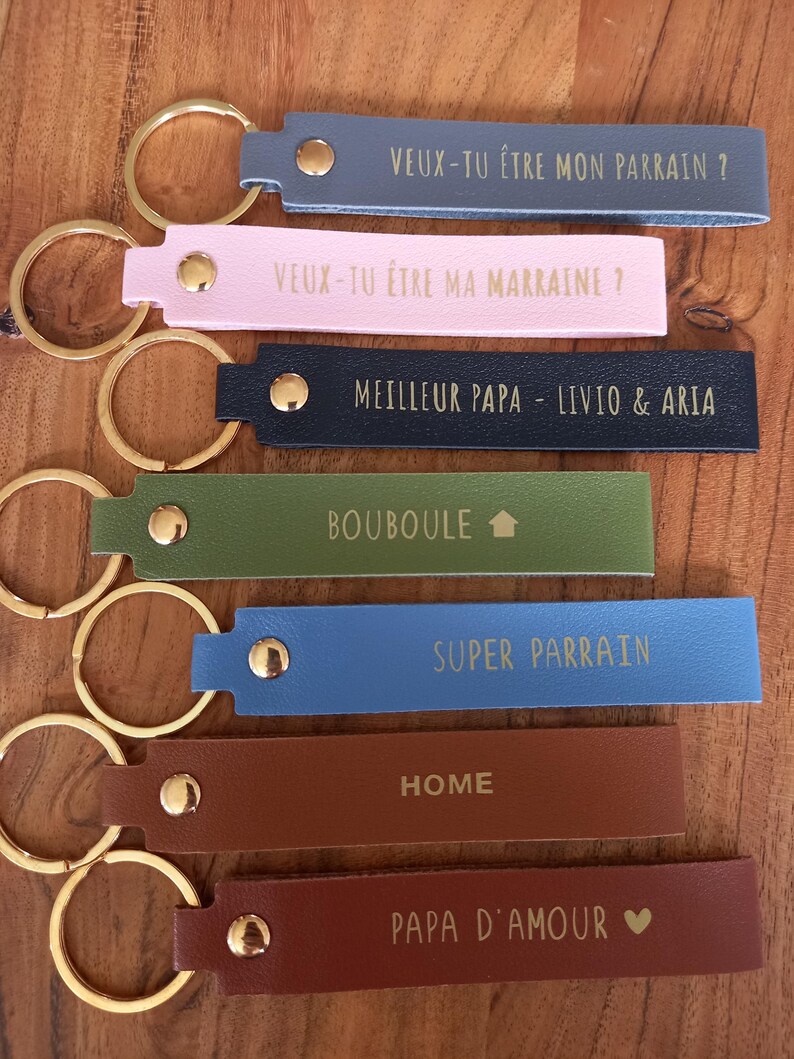 May include: Seven rectangular keychains in various colours including blue, pink, navy, green, light blue, and brown. Each features a gold metal ring, a gold rivet, and gold text including phrases like Veux-tu &ecirc;tre mon parrain, Meilleur Papa, and Home.