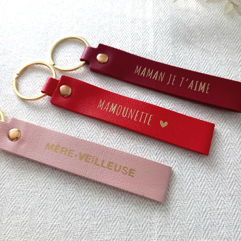 May include: Three rectangular keychains in burgundy, red, and light pink, each featuring a gold metal ring and a gold rivet. The burgundy keychain is embossed with MAMAN JE T'AIME, the red one with MAMOUNETTE and a heart, and the pink one with M&Egrave;RE-VEILLEUSE.