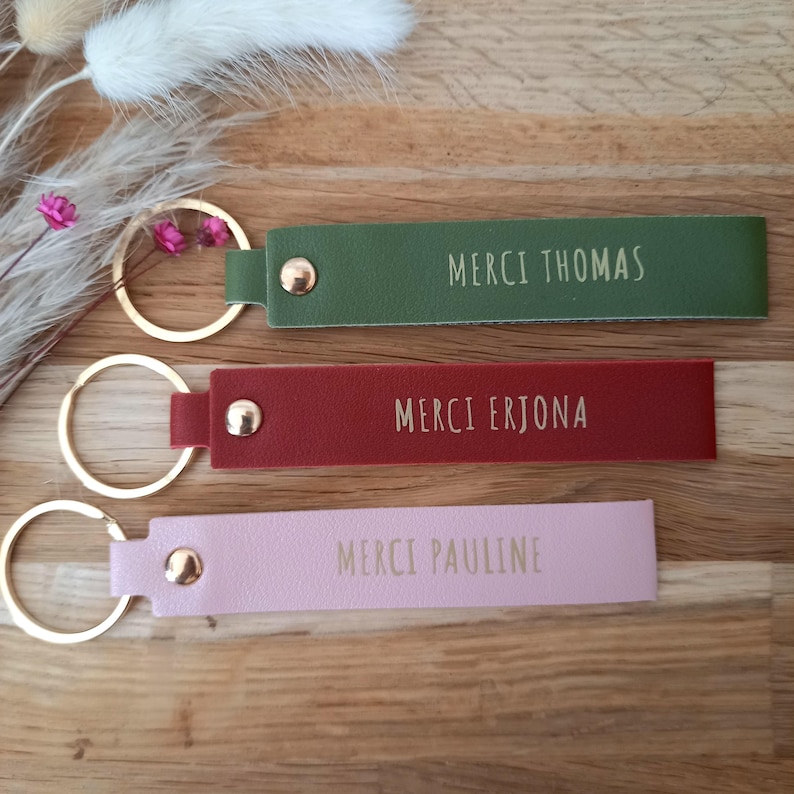 May include: Three rectangular keychains in olive green, dark red, and light pink, each featuring a gold-tone metal ring and a gold-tone rivet. The keychains are embossed with the text MERCI THOMAS, MERCI ERJONA, and MERCI PAULINE respectively.