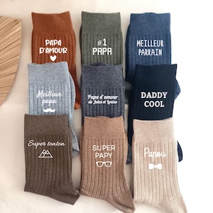 Puede incluir: Una colección de nueve calcetines acanalados en varios colores, incluyendo óxido, oliva, azul, gris, carbón, negro, marrón, tostado y beige. Cada calcetín presenta texto e iconos impresos en blanco como Papa d'amour, #1 Papa, Meilleur Parrain, Daddy Cool, Super tonton, Super Papy y Papou.