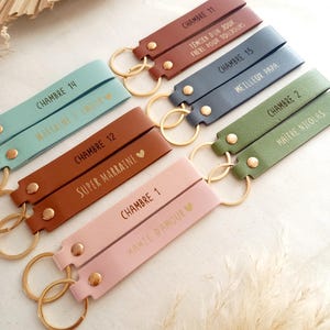 May include: A collection of rectangular leather keychains in various colours including light blue, brown, dark blue, green, and pink. Each keychain features a gold metal ring, a gold rivet, and custom text such as CHAMBRE and names or titles.