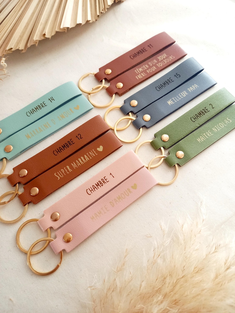 May include: A collection of rectangular leather keychains in various colours including light blue, brown, dark blue, green, and pink. Each keychain features a gold metal ring, a gold rivet, and custom text such as CHAMBRE and names or titles.