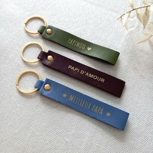 May include: Three rectangular keychains with gold-tone metal rings and rivets. The top is olive green with the text PAPINOU and a heart. The middle is dark purple with the text PAPI D'AMOUR. The bottom is light blue with the text MEILLEUR PAPA and stars.