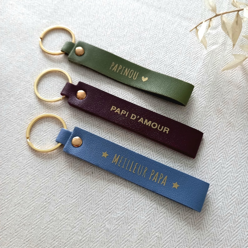 May include: Three rectangular keychains with gold-tone metal rings and rivets. The top is olive green with the text PAPINOU and a heart. The middle is dark purple with the text PAPI D'AMOUR. The bottom is light blue with the text MEILLEUR PAPA and stars.