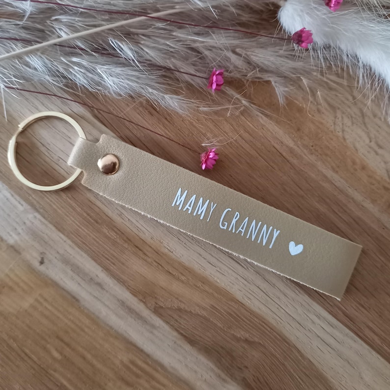 May include: A beige leather keyring featuring a gold-toned metal ring and a gold rivet. The strap is printed with the white text MAMY GRANNY followed by a small white heart symbol. The keyring has a smooth texture and a rectangular shape.
