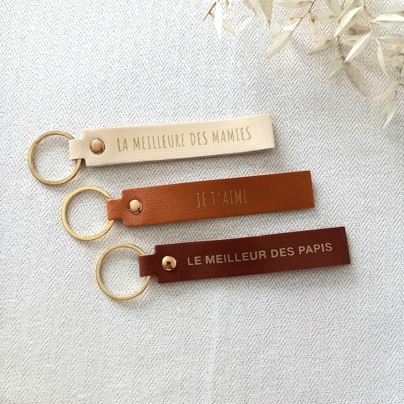 May include: Three rectangular leather keychains with gold-tone metal rings and rivets. The top keychain is cream with the text LA MEILLEURE DES MAMIES, the middle is tan with JE T'AIME, and the bottom is dark brown with LE MEILLEUR DES PAPIS.