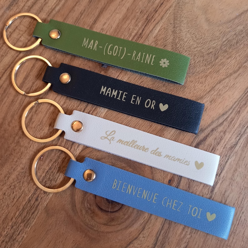 May include: Four rectangular keychains with gold-tone metal rings and rivets. The olive green one reads MAR-(GOT)-RAINE with a flower, the navy blue reads MAMIE EN OR with a heart, the white reads La meilleure des mamies with a heart, and the blue reads BIENVENUE CHEZ TOI with a heart.