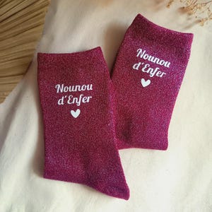 Peut inclure: Une paire de chaussettes magenta pailletées avec le texte blanc Nounou d'Enfer et une petite icône de cœur blanc imprimés sur la partie supérieure de chaque chaussette. Le tissu présente une finition métallique scintillante sur toute la surface.