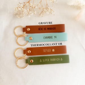 May include: Four rectangular leather keychains with gold-tone metal rings and rivets. The top two are labelled GRAVURE, featuring brown and light blue colours with text R&Ecirc;VE DE MARKHOR and CHAMBRE 14. The bottom two are labelled THERMOCOLLANT OR, in rust and olive green with text PAPHAD and SUPER PARRAIN.