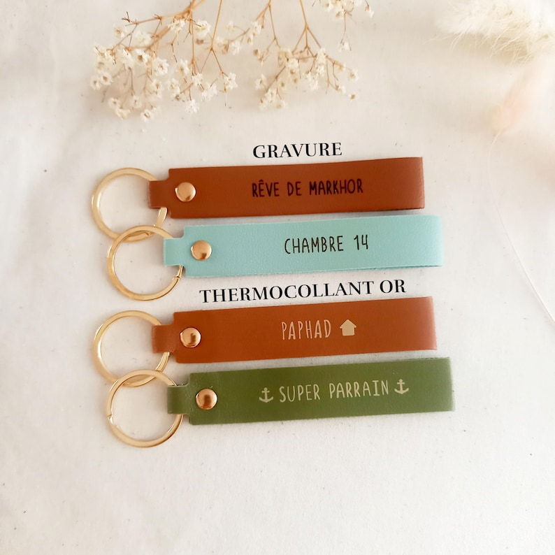 May include: Four rectangular leather keychains with gold-tone metal rings and rivets. The top two are labelled GRAVURE, featuring brown and light blue colours with text R&Ecirc;VE DE MARKHOR and CHAMBRE 14. The bottom two are labelled THERMOCOLLANT OR, in rust and olive green with text PAPHAD and SUPER PARRAIN.