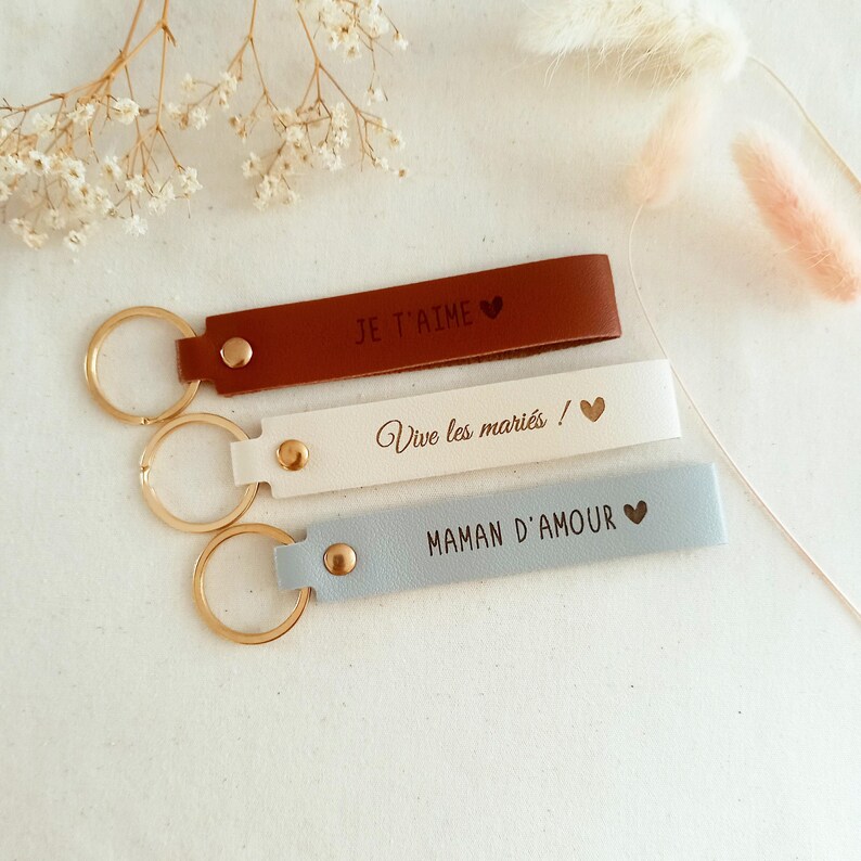 May include: Three rectangular leather keychains with gold-tone metal rings and rivets. The top keychain is brown with the text JE T'AIME and a heart. The middle is cream with Vive les mari&eacute;s ! and a heart. The bottom is light blue with MAMAN D'AMOUR and a heart.